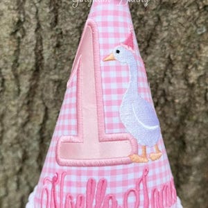 May include: Pink and white gingham fabric birthday hat with a white pom pom on top. The hat has a pink number one and a white goose with a pink hat embroidered on it. The name "Noella Jane" is embroidered on the bottom of the hat.