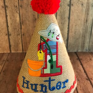May include: A burlap cone-shaped party hat with a red pom-pom, a fishing-themed design, and the name "Hunter" embroidered in blue. The design includes a fishing rod, a fish, and a bucket. The hat is trimmed with red ric-rac.