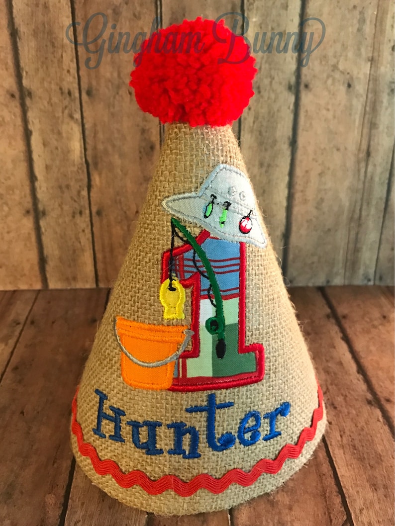 May include: A burlap cone-shaped party hat with a red pom-pom, a fishing-themed design, and the name "Hunter" embroidered in blue. The design includes a fishing rod, a fish, and a bucket. The hat is trimmed with red ric-rac.