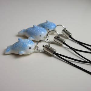 May include: Three small whale shark charms made of a smooth, light blue material with white polka dots. Each charm features a black eye and is attached to a silver-toned metal jump ring connected to a thin, black cord strap for attachment.