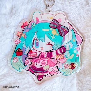 Puede incluir: Un llavero acrílico transparente con un personaje de dibujos animados con cabello turquesa, un vestido rosa y una diadema con orejas de conejo. El llavero tiene un cierre plateado en forma de estrella y está decorado con corazones, estrellas y un pastel pequeño.