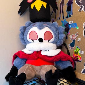 May include: A plush toy featuring a blue, furry creature with large red eyes and a black headpiece with yellow and pink accents. The toy has a red cape, white collar, and brown pants. Black feet and arms complete the design. The toy is sitting on a black metal shelf.