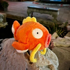 May include: An orange plush toy fish with white and yellow details. The toy has a large, round white eye with a black pupil, and a red mouth. The fish is resting on a grey rock.