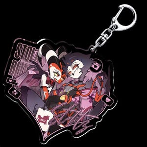 May include: A black and white acrylic keychain featuring two cartoon characters, one red and one purple, with the text "Stolas Blitz" and "Stolas" in pink.