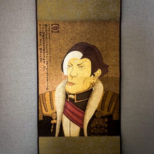 May include: A vertical scroll painting depicting a person in ornate clothing. The artwork uses gold, brown, and red hues, with detailed imagery and text. The scroll is suspended by a brown cord and has a black roller at the bottom.