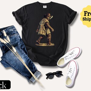May include: Black t-shirt featuring a Bigfoot graphic wearing a poncho and cowboy hat. The image includes blue jeans, white sneakers, and black sunglasses. The word "Black" is displayed on a black rectangle.