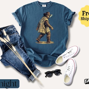 May include: A navy blue t-shirt featuring a Bigfoot graphic wearing a poncho and hat. The image includes a pair of blue jeans, white sneakers, and black sunglasses. The word "Midnight" is displayed on the bottom left.