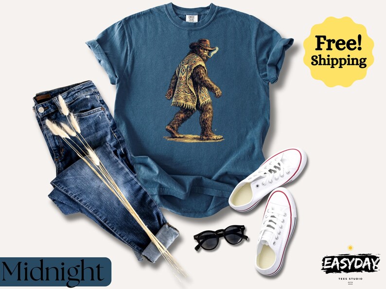 May include: A navy blue t-shirt featuring a Bigfoot graphic wearing a poncho and hat. The image includes a pair of blue jeans, white sneakers, and black sunglasses. The word "Midnight" is displayed on the bottom left.