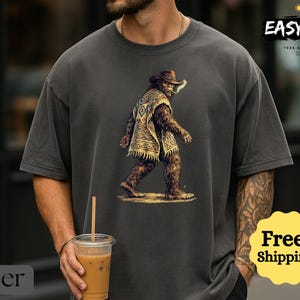 May include: Dark gray t-shirt featuring a Bigfoot illustration wearing a cowboy hat and poncho. The image includes the text "EASYDAY" and "Free! Shipping". The shirt is a relaxed fit.