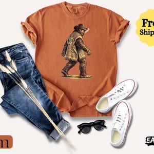 May include: An orange t-shirt with a Bigfoot graphic, paired with blue jeans, white sneakers, and black sunglasses. The shirt features a Bigfoot illustration wearing a hat and poncho. The word "Yam" is at the bottom left.