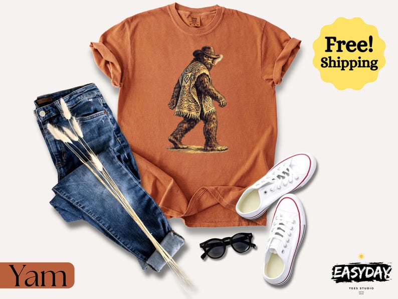 May include: An orange t-shirt with a Bigfoot graphic, paired with blue jeans, white sneakers, and black sunglasses. The shirt features a Bigfoot illustration wearing a hat and poncho. The word "Yam" is at the bottom left.