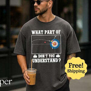 May include: Dark grey t-shirt with the text "WHAT PART OF DON'T YOU UNDERSTAND?" and a curling diagram. The shirt features a graphic of a curling target and stones. The person is holding a beverage in a clear plastic cup.