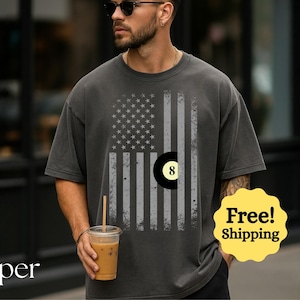 May include: Dark gray t-shirt featuring a distressed American flag design with an eight-ball graphic. The shirt has a relaxed fit and short sleeves. The text "Peper" is at the bottom left.