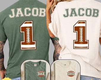 May include: A sage green t-shirt and a cream t-shirt, each featuring the name JACOB and the number 1 in a football-themed graphic on the back. The front of each shirt displays a small football graphic with the text DAD or MOM respectively.