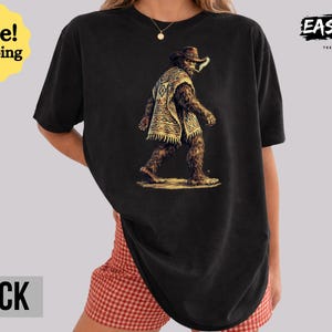May include: Black t-shirt featuring a Bigfoot illustration wearing a cowboy hat and poncho. The graphic depicts the creature walking with a cigarette in its mouth. The shirt has the text "BLACK" at the bottom.