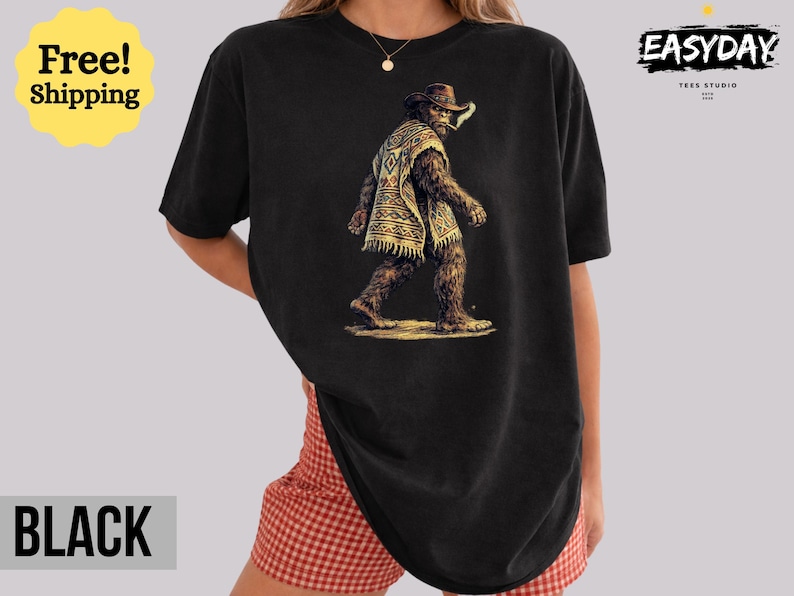 May include: Black t-shirt featuring a Bigfoot illustration wearing a cowboy hat and poncho. The graphic depicts the creature walking with a cigarette in its mouth. The shirt has the text "BLACK" at the bottom.