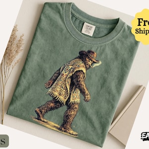 May include: Moss green t-shirt featuring a Bigfoot illustration wearing a cowboy hat and poncho. The graphic depicts Bigfoot smoking a cigarette. The shirt has a crew neck and short sleeves. The text "Comfort Colors" is visible on the tag.