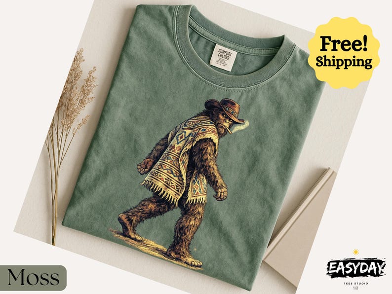 May include: Moss green t-shirt featuring a Bigfoot illustration wearing a cowboy hat and poncho. The graphic depicts Bigfoot smoking a cigarette. The shirt has a crew neck and short sleeves. The text "Comfort Colors" is visible on the tag.
