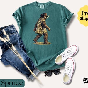 May include: A teal t-shirt with a graphic of a Bigfoot-like creature wearing a hat and poncho, paired with blue jeans, white sneakers, and black sunglasses. The words "Blue Spruce" are at the bottom.