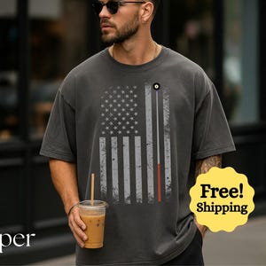 May include: Dark gray t-shirt featuring a distressed American flag design with a pool cue and ball. The shirt has a relaxed fit and short sleeves. The person in the image is holding a cup of iced coffee.
