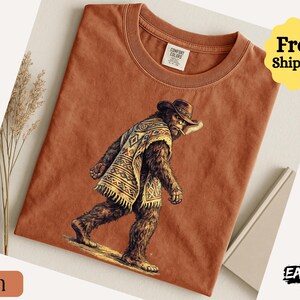 May include: A terracotta-colored t-shirt featuring a Bigfoot-like creature wearing a cowboy hat and poncho. The graphic is detailed with brown, tan, and cream colors. The shirt has a crew neck and the Comfort Colors label is visible.