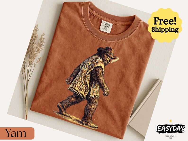 May include: A terracotta-colored t-shirt featuring a Bigfoot-like creature wearing a cowboy hat and poncho. The graphic is detailed with brown, tan, and cream colors. The shirt has a crew neck and the Comfort Colors label is visible.