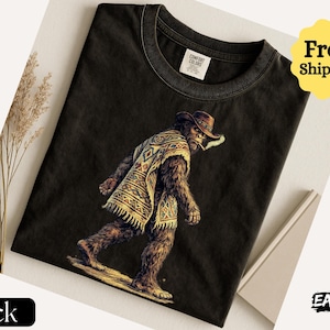 May include: Black t-shirt featuring a Bigfoot character wearing a cowboy hat and poncho, smoking a cigarette. The graphic is detailed with brown, tan, and yellow colors. The shirt has a Comfort Colors tag. The words "Black" and "EASYDAY" are also visible.