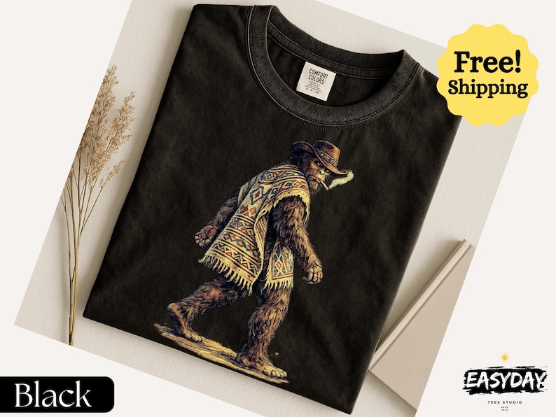 May include: Black t-shirt featuring a Bigfoot character wearing a cowboy hat and poncho, smoking a cigarette. The graphic is detailed with brown, tan, and yellow colors. The shirt has a Comfort Colors tag. The words "Black" and "EASYDAY" are also visible.