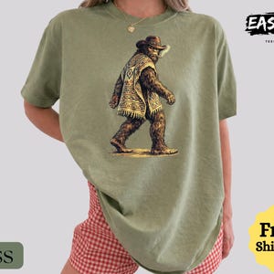 May include: Moss green t-shirt featuring a graphic of Bigfoot wearing a cowboy hat and poncho. The shirt has the text "EASYDAY TEES STUDIO" in the upper right corner. The word "Moss" is in a green rectangle in the lower left corner.