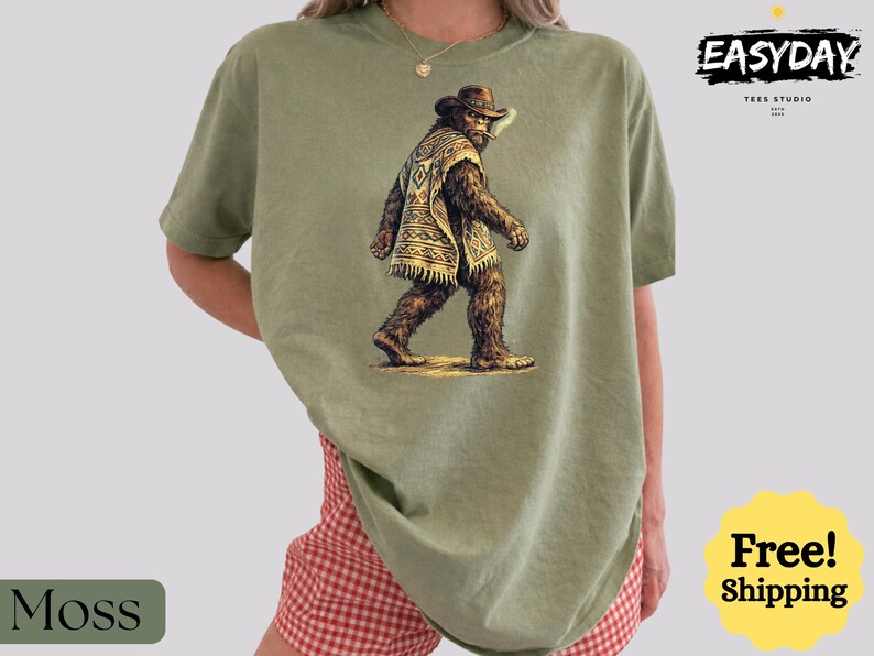 May include: Moss green t-shirt featuring a graphic of Bigfoot wearing a cowboy hat and poncho. The shirt has the text "EASYDAY TEES STUDIO" in the upper right corner. The word "Moss" is in a green rectangle in the lower left corner.