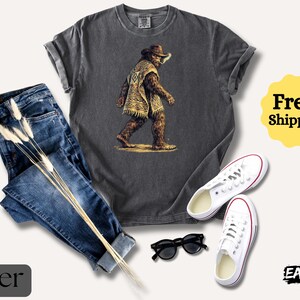 May include: A charcoal gray t-shirt featuring a Bigfoot illustration in a cowboy hat and poncho. The image is paired with blue jeans, white sneakers, and black sunglasses. The words "Free! Shipping" are in a yellow circle.