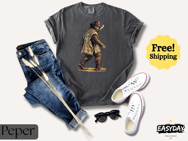 May include: A charcoal gray t-shirt featuring a Bigfoot illustration in a cowboy hat and poncho. The image is paired with blue jeans, white sneakers, and black sunglasses. The words "Free! Shipping" are in a yellow circle.