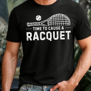 May include: Black t-shirt with a white graphic of a tennis racket and ball. The text "TIME TO CAUSE A RACQUET" is printed below the racket. The shirt is made of a soft material.
