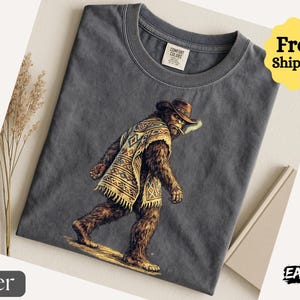 May include: Dark gray t-shirt featuring a Bigfoot illustration wearing a cowboy hat and poncho, smoking a cigarette. The shirt has a crew neck and a tag that says "Comfort Colors". The image also includes the text "Free! Shipping".