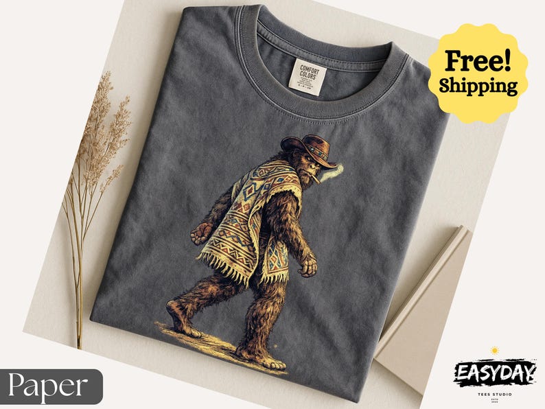 May include: Dark gray t-shirt featuring a Bigfoot illustration wearing a cowboy hat and poncho, smoking a cigarette. The shirt has a crew neck and a tag that says "Comfort Colors". The image also includes the text "Free! Shipping".