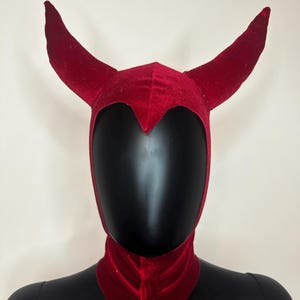 May include: A deep red velvet hood featuring two prominent, curved horns extending from the top. The headpiece covers the crown and neck area, with a pointed front detail above the forehead and a structured opening for the face.
