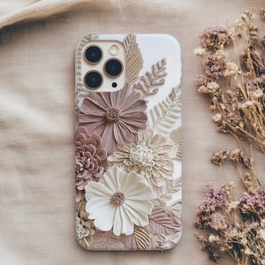 May include: A white smartphone case featuring a three-dimensional floral design with textured petals in shades of dusty rose, cream, and beige. The arrangement includes various sculpted flowers and fern-like leaves covering the back surface.