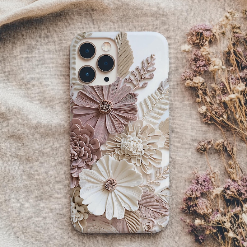 May include: A white smartphone case featuring a three-dimensional floral design with textured petals in shades of dusty rose, cream, and beige. The arrangement includes various sculpted flowers and fern-like leaves covering the back surface.