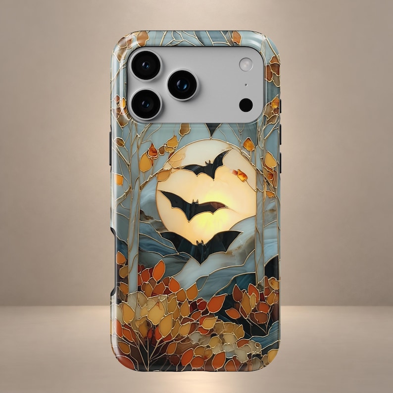 May include: A smartphone case featuring a stained glass style design of three black bats flying in front of a large, glowing yellow moon. The scene includes stylized trees and autumn-colored orange and brown foliage, all outlined in gold lines.