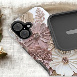 May include: A protective phone case featuring a raised, textured floral design in shades of dusty rose, cream, and beige. The case has a black camera cutout frame and a black interior lining, showcasing detailed petals and fern-like botanical elements.