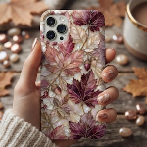 May include: A smartphone case featuring a detailed pattern of autumn leaves in shades of deep burgundy, soft pink, and cream, accented with small brown acorns. The design has a pearlescent, iridescent finish that catches the light across the surface.