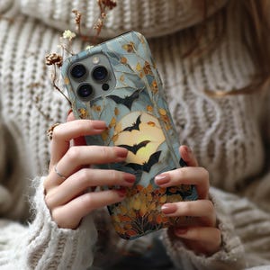 May include: A smartphone case featuring a stained glass design with a pale yellow full moon, black bat silhouettes, and autumn-colored foliage in shades of orange and brown, all outlined in gold against a light blue background.