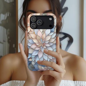 Puede incluir: Funda de teléfono con un diseño floral en colores pastel. La funda tiene un módulo de cámara negro y se sostiene frente a la cara de una persona. El diseño floral incluye tonos de rosa, azul y beige.