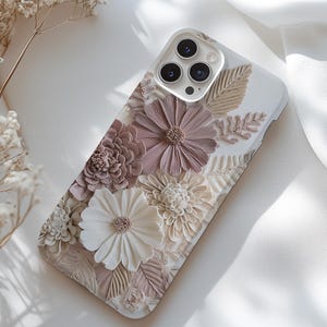 Könnte beinhalten: Weißes Handycase mit 3D-Blumendesign. Das Case zeigt verschiedene Blumen in Weiß-, Beige- und Mauve-Tönen mit detaillierten Blütenblattstrukturen und Blättern. Die Kameralinsen des Telefons sind oben sichtbar.