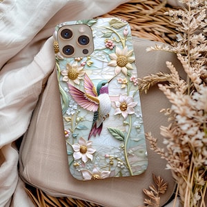 May include: A white phone case with a 3D design of a hummingbird flying over flowers. The flowers are pink, white, and gold. The phone case has a camera lens cutout.