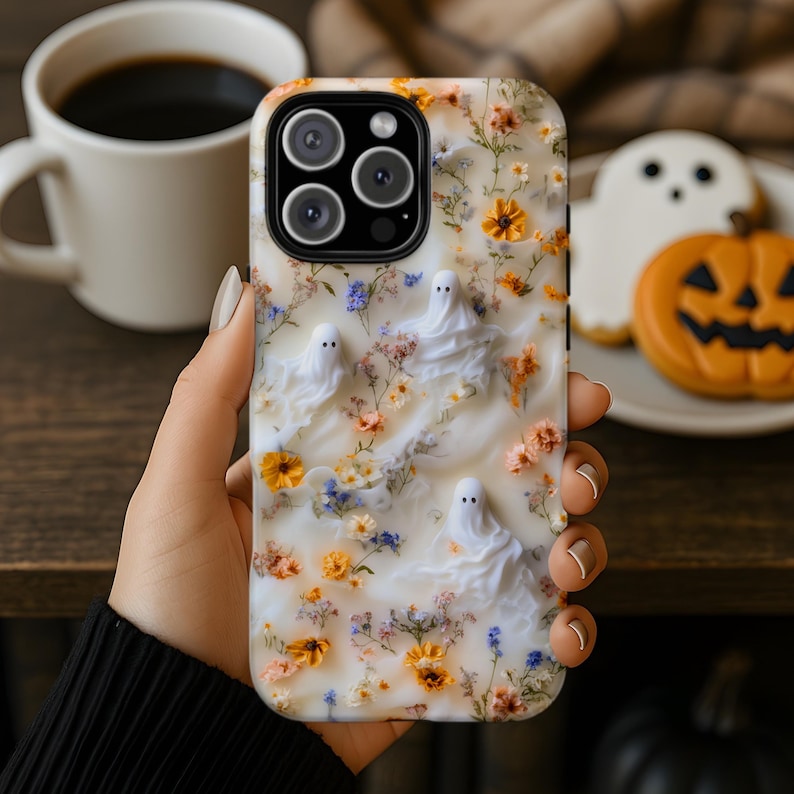 Puede incluir: Una funda para tel&eacute;fono con fondo blanco marmoleado decorado con peque&ntilde;as flores prensadas de color naranja, amarillo y azul. Tres peque&ntilde;as figuras blancas con forma de fantasma est&aacute;n dispersas por la superficie, integradas en el dise&ntilde;o floral.