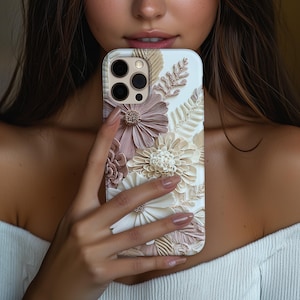 May include: A smartphone case featuring a three-dimensional floral design with layered petals in shades of cream, beige, and dusty rose. The surface is adorned with intricate sculpted flowers and fern-like leaves, creating a textured relief pattern.