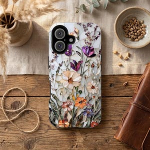 Puede incluir: Funda de teléfono con diseño floral de mosaico, con flores blancas, moradas, naranjas y beige. La funda tiene un recorte para una cámara de doble lente. Colocada sobre una superficie de madera con elementos decorativos.