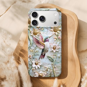 Peut inclure: Coque de téléphone ornée d'un motif de colibri en 3D, avec des plumes roses, violettes et vertes, entouré de fleurs blanches et dorées. Le fond est bleu clair et blanc, avec une découpe pour l'appareil photo.
