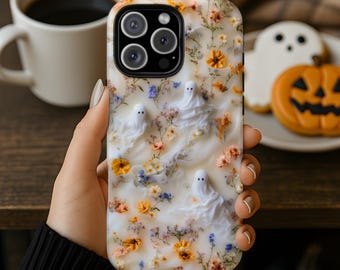 May include: A smartphone case with a white, marbled background featuring embedded dried orange and blue flowers and three small, white, raised ghost figures. The case has a black camera cutout frame and a glossy finish over the floral design.